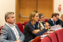 Grupa osób w średnim wieku, siedzą w auli na czerwonych fotelach. Na stolikach przed nimi leżą papiery. Jedna z osób, kobieta, mówi do trzymanego w ręce mikrofonu. Patrzą przed siebie.