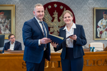 Mężczyzna i kobieta, elegancko ubrani, pozują do zdjęcia, uśmiechając się. Kobieta trzyma w dłoniach otwartą teczkę.