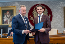 Dwóch mężczyzn, elegancko ubranych, pozuje do zdjęcia, uśmiechają się. Mężczyzna po prawej trzyma w rękach teczkę.