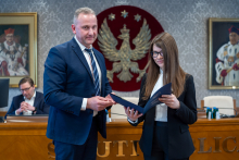 Mężczyzna i kobieta, elegancko ubrani, pozują do zdjęcia, uśmiechając się. Kobieta trzyma w dłoniach otwartą teczkę.