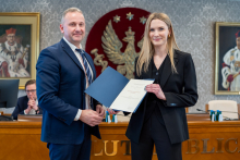 Mężczyzna i kobieta, elegancko ubrani, pozują do zdjęcia, uśmiechając się. Kobieta trzyma w dłoniach otwartą teczkę.