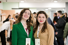 Dwie młode kobiety, organizatorki wydarzenia. Obie są elegancko ubrane. Uśmiechają się do zdjęcia. Kobieta po lewej ma zieloną marynarkę i identyfikator zawieszony na szyi. Kobieta po prawej ma beżową bluzkę i identyfikator zawieszony na szyi. W tle znajduje się grupa ludzi.