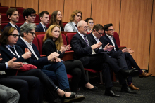 Uczestnicy konferencji. Osoby w różnym wieku, mężczyźni i kobiety. Siedzą na czerwonych fotelach i patrzą na wprost. Są elegancko ubrani. Biją brawo, uśmiechają się.