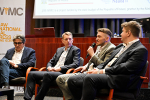 Czterech mężczyzn. Biorą udział w panelu dyskusyjnym. Siedzą w auli w fotelach. Są ubrani w garnitury. Jeden z nich trzyma mikrofon i przemawia. W tle widoczny jest ekran projekcyjny z logo "WIMC".