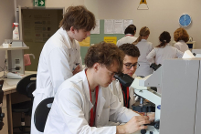 Wnętrze laboratorium. Na pierwszym planie znajduje się mężczyzna w białym fartuchu laboratoryjnym, który patrzy przez mikroskop. Za nim stoi dwóch innych mężczyzn, również w białych fartuchach. Są skupieni na rozwiązywaniu zadania. To nasi studenci. W tle widocznych jest kilka innych osób, które wykonują różne czynności laboratoryjne. Na tylnej ścianie zawieszone są dokumenty oraz zegar.