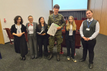 Sześć osób stoi w rzędzie w sali konferencyjnej. Są to studenci i organizatorzy konkursu. Trzy osoby po lewej stronie są ubrane formalnie. Osoba w środku nosi mundur wojskowy i trzyma teczkę z dokumentami. Dwie osoby po prawej stronie są ubrane formalnie i mają identyfikatory na szyjach. W tle widać ekran projekcyjny i drewniane panele ścienne.