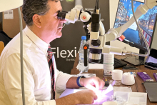 The man, leading the workshop, is sitting at a desk with a microscope on it and looking through it. He is wearing a white shirt and tie. There are various objects on the desk.