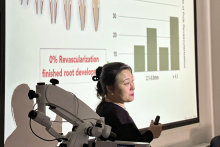 A woman conducting a workshop. On the left, next to her, you can see a microscope. Behind her is a large multimedia screen. The screen displays images of teeth and a bar graph.