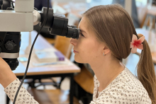 A young woman, a workshop participant. She is dressed in a light blouse and has long hair. She is looking through a microscope.