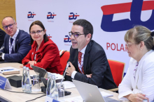 Cztery osoby siedzą przy stole konferencyjnym. Mężczyźni mają na sobie granatowe marynarki i białe koszule. Mężczyzna siedzący jako drugi z prawej strony mówi do mikrofonu. Pierwsza z prawej siedzi kobieta w białej marynarce. Patrzy przed siebie. Od lewej strony siedzą mężczyzna i kobieta. Kobieta ma na sobie czerwony żakiet. Oboje patrzą na mówiącego. Na stole znajdują się mikrofony, butelki z wodą, filiżanki oraz tabliczki z nazwiskami.