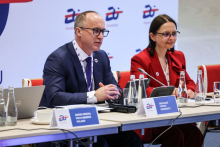 Kobieta i mężczyzna siedzą przy stole.  Mężczyzna po lewej stronie ma na sobie ciemny garnitur i białą koszulę, a kobieta po prawej stronie ma na sobie czerwony strój. Na stole znajdują się mikrofony, butelki z wodą, filiżanki oraz tabliczki z nazwiskami.