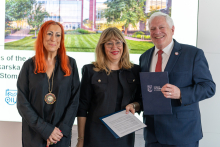 The group consists of three people: two women - the organizers of the event and a man - one of the speakers. They are elegantly dressed and smiling at the camera. In the background is a large screen displaying text and an image of a building surrounded by greenery. - One of the speakers (right). They are elegantly dressed and smiling at the camera. In the background is a large screen displaying text and an image of a building surrounded by greenery. 