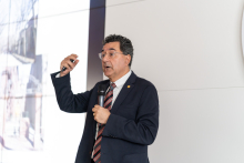 One of the conference speakers and presenter of the endodontics workshop. A middle-aged man, wearing a suit and glasses. He holds a microphone in one hand and has the other raised and gestures with it. He is speaking. A screen with a projected image is visible in the background.