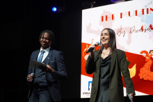 Young people standing on stage. The man on the left wears a dark suit. He holds a microphone and a piece of paper. The woman on the right wears a black dress and a green jacket. She is also holding a microphone. Behind them is a large screen. 