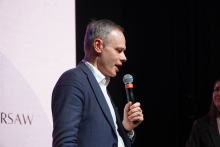 A middle-aged man. He wears a navy blue jacket and a white shirt. He holds a microphone to his mouth. The background is dark.