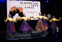 Five female dancers performing a traditional Korean drum dance. They wear yellow blouses and purple skirts. Each holds a drum and drumsticks. A large screen can be seen in the background. The screen reads: “TRADITIONAL KOREAN DRUM DANCE.” Speakers and technical equipment stand on the stage. In front of the stage the audience is sitting. One of the spectators takes a photo or records with his phone.