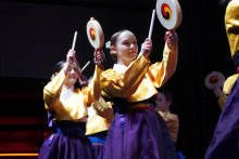A group of young women dancing on stage. They wear yellow shirts and purple skirts. Each person holds a round drum. On the drum is a colorful pattern in the shape of a spiral. The spiral has the colors red, blue and yellow. The background is dark. The stage is brightly lit.