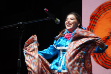 A woman standing on stage behind a microphone. She has dark hair. She smiles and sings. She wears a blue outfit with white decorations. She has a pink scarf around her neck. She holds a colorful skirt and spreads it out to the sides. The background is dark. A large orange pattern can be seen on the right side. 