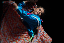 A dancing woman. She has dark hair, braided into pinned up braids. She wears a blue blouse. She has a colorful skirt with a floral pattern. The background is black. 