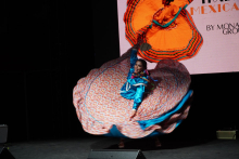 Dancing woman. The woman has dark hair. She wears a blue blouse. She has a colorful skirt with a floral pattern. The background is black. 