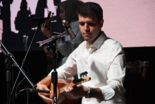 Two men on stage. The first man sits on a chair. He wears a white shirt. A microphone stands in front of him. The second man stands at the back. He is also holding a guitar. There is also a microphone in front of him. The background is dark. In the background you can see light silhouettes of figures.