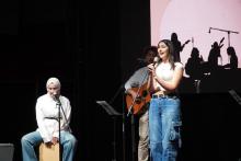 Three young people standing and sitting on stage. The woman on the left is sitting on a cajon. She wears a white hoodie and blue jeans. The woman in the middle stands and sings. She is wearing a light-colored shirt and beige pants. The person on the right stands at the microphone. She wears a white sleeveless shirt and blue jeans with pockets. A large screen can be seen in the background. On the screen are black silhouettes of musicians with instruments.