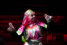 A woman dancing on stage. She wears a traditional colorful folk costume. She has a headscarf on her head in pink and yellow. The upper part of the costume is pink. It has white and gold decorations. There are green accents on the sleeves. The person raises his hands. She looks as if she is dancing or making gestures. The background is dark. Red lights can be seen in the background.