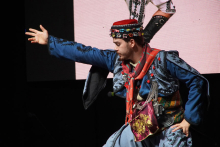 A man in traditional folk costume. The costume is blue with black patterns and red accents. The man is wearing a red cap with colorful decorations on his head. The right hand is stretched forward. The left hand rests on the hip. A large screen can be seen in the background.