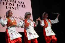 Three women dancing a traditional Turkish folk dance. They wear white shirts and red pants. They have white aprons with colorful embroidery. On their heads are headscarves. A large screen can be seen in the background. The screen reads: “TRADITIONAL TURKISH FOLK DANCE”. 