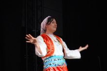 A woman on stage. She is wearing traditional folk costume. She wears a white long-sleeved shirt, a red vest with embroidery and a blue belt decorated with gold coins. On his head is a white scarf. The background is dark. On the left you can see the metal structure of the stage.