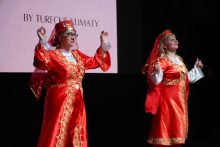 Two women dressed in traditional Turkish dance costumes. The costumes are red and have rich gold decorations. Both wear red headscarves on their heads. Their hands are raised in a dance gesture. In the background is a screen with the words “BY TURKISH CLIMATES.”