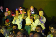 A group of people standing and clapping. In the background is a DJ at a console. Above him shine colored lights - you can see shades of blue, pink and purple. The faces of the people in the foreground are blurred. Some are wearing traditional outfits, while others are dressed in casual clothes. Dark colors dominate, but stage lights add bright accents.