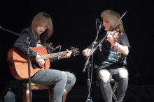 Dwie osoby grające na instrumentach muzycznych. Osoba po lewej stronie gra na gitarze akustycznej. Osoba po prawej stronie gra na skrzypcach. Obie siedzą na krzesłach. Mają długie włosy, które zasłaniają im twarze. Tło jest ciemne. Światło pada na osoby i ich instrumenty.