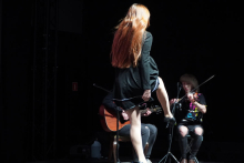 A woman with long red hair dancing in the middle of the stage. She is dressed in a black dress. She has one leg raised. Behind her sits two people. One is playing an acoustic guitar, the other a violin. The background is dark. The stage is lit with a bright light.