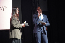 A young woman and a young man standing on stage. The woman on the left has long, dark hair. She wears a beige jacket and holds a microphone in her right hand. The man on the right is wearing a navy blue suit and tie. He holds a microphone in his left hand and a piece of paper in his right hand. The background is dark. A portion of the screen can be seen on the left.