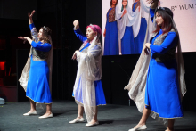 Three young women dancing on stage. They are wearing blue dresses with long sleeves. On top of the dresses are gold patterns. On their shoulders are transparent gold scarves. The dancers stand in various poses with their arms raised. The background is dark. Behind them is a large screen with a picture of other people in similar costumes.