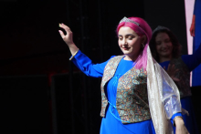 A young woman dressed in a blue dress and a patterned vest. One hand is raised in the air. The woman has pink hair. 