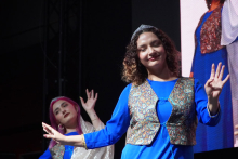 Two young women dancing on stage. The woman in the foreground has curly hair. She wears a blue dress and a patterned vest. In the background, a second person in a similar outfit can be seen. 