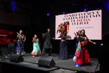 A group of people dancing on stage. They are wearing traditional Indian costumes in different colors - blue, green, red and black. In the background is a large screen with the words “Contemporary & modern elements of Indian culture by WUM students.” The stage is illuminated with different colored lights.