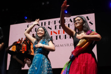 Two women in the foreground, man in the background on the left. The woman on the left wears a blue dress with white patterns. The woman on the right is wearing a black blouse, red skirt and a colorful shawl. In the background is a large screen with the caption “Contemporary Indian elements in culture”.