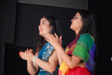 Two women standing side by side. The woman on the left wears a blue dress with white patterns and a silver bracelet on her wrist. The woman on the right has a colorful scarf in rainbow shades and a red skirt. Both have long, dark hair and are clapping their hands. The background is dark, with a black wall or curtain behind them.