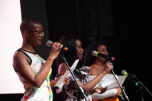 Three women standing on stage. Each holds a microphone and a piece of paper. The woman on the left has short hair and wears a colorful sleeveless shirt. The woman in the middle has long, dark hair and wears a dress with a pattern. The woman on the right has curly hair. Also she is holding a microphone and a piece of paper. The background is dark. The people are illuminated by the stage light.