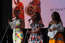 Three people standing on the stage. Each holds a piece of paper and stands by a microphone. The woman on the left is wearing a colorful dress. The woman in the middle has a dress with a circle pattern and frills. The person on the right has a white long-sleeved blouse and a yellow skirt with blue patterns. There is a large screen in the background. The screen shows two people in traditional African costumes and the caption.