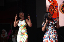 Two women standing on stage. The woman on the left wears a tight dress with colorful patterns - white, yellow, green and blue. The woman on the right is wearing a short-sleeved dress with white, blue and red patterns. Both people have their hands raised and are pointing their fingers upward. In the background is a large screen with a picture of a person in a red dress with black and gold elements. 