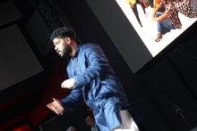 A man in a blue shirt standing on a stage. His face is blurred. There is a large screen in the background. The screen displays a photo of a group of people sitting on the floor. The scene is dark. The light falls on the man.