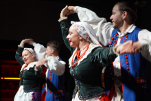 Four people dancing in traditional folk costumes. Two women wear green blouses with white sleeves, red beads and white scarves on their heads. Two men have blue vests with red buttons and white shirts. They all hold hands and raise them up. The background is dark and the figures are well lit.