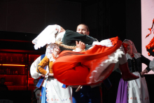 A group of people in traditional folk costumes. One person lifts another. The person being lifted has a white scarf on his head and a red skirt with white laces. The background is a stage illuminated by a red light. 