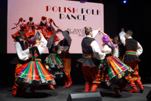 A group of dancers performing a Polish folk dance. They wear traditional folk costumes. The costumes consist of colorful skirts, vests and red shoes. In the background is a large screen with the words “POLISH FOLK DANCE”.