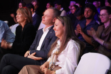 A group of people sitting on white chairs. In the foreground sits a man in a dark suit and a woman in a white shirt with long hair. Other people can be seen in the background, some clapping. The background is dark. The stage lighting creates colorful reflections.