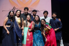 Nine male and female WUM students stand on a stage. Behind them is a large screen with a “thank you” sign. They are wearing colorful traditional clothes in shades of blue, red, green and gold. They smile and pose for a photo.
